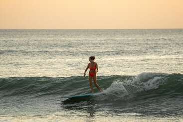 A woman in a red swimsuit surfs on a small wave during sunset, balancing confidently on her surfboard with the calm ocean stretching out behind her.