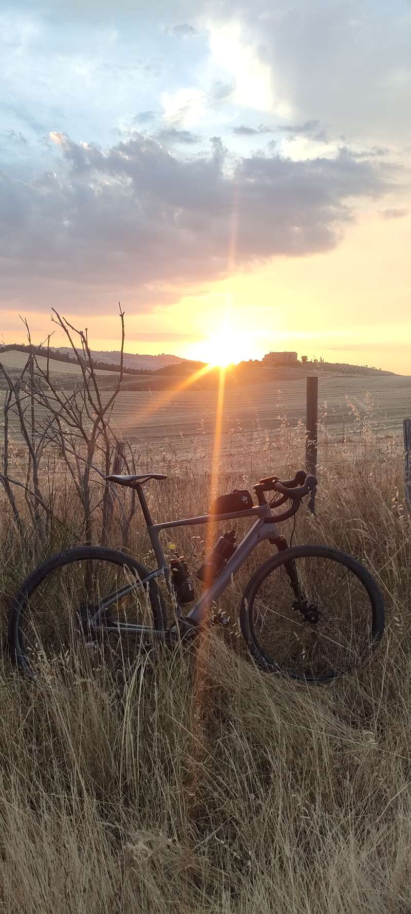 Uma bicicleta descansa na grama alta e dourada ao lado de uma cerca, enquanto o sol se põe sobre colinas distantes e uma casa, lançando uma luz quente e dramática.