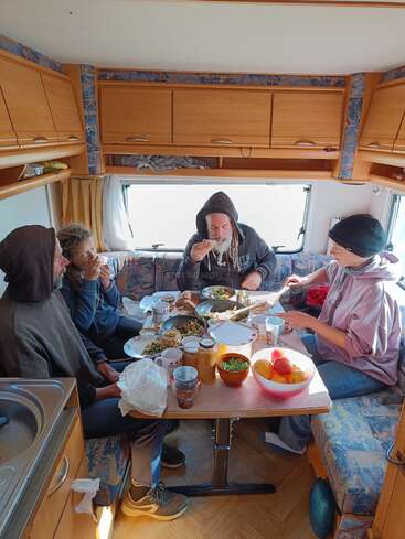 L'image représente quatre personnes assises autour d'une table dans un camping-car, en train de prendre un repas. La table est chargée de nourriture et de boissons.