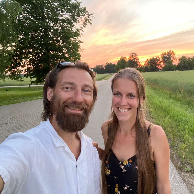 Um casal sorridente está em uma estrada rural ao pôr do sol. Atrás deles, dá pra ver campos verdes e árvores. O céu está lindamente iluminado com cores quentes.
