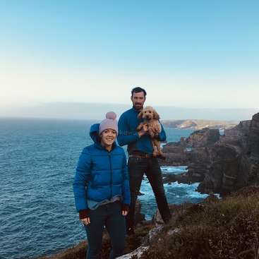 L'image représente un homme et une femme debout sur une falaise surplombant l'océan, l'homme tenant un petit chien. Le couple est habillé chaudement.