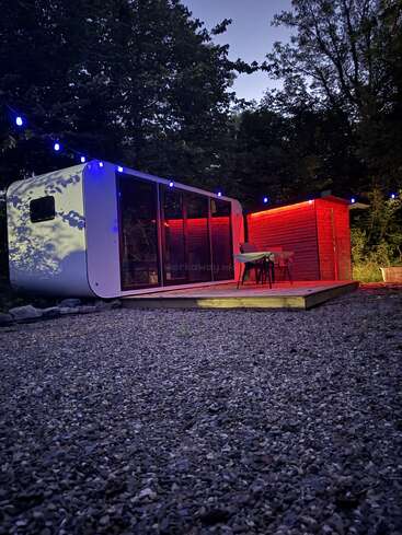 Une petite maison moderne avec des portes en verre et des lumières rouges brillantes est assise sur une terrasse en bois, entourée d'arbres et de guirlandes lumineuses bleues au crépuscule. Un cadre paisible.