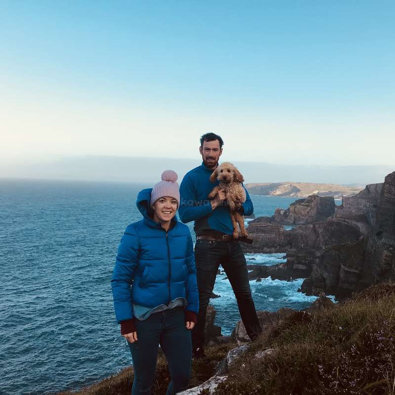 L'image représente un homme et une femme debout sur une falaise surplombant l'océan, l'homme tenant un petit chien. Le couple est habillé chaudement.