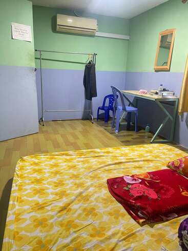 Cette chambre sobre est équipée d'un matelas recouvert d'une parure de lit à motifs floraux jaunes, d'une couverture rouge, d'un bureau avec une chaise, d'un miroir, d'un porte-manteau et d'un climatiseur.