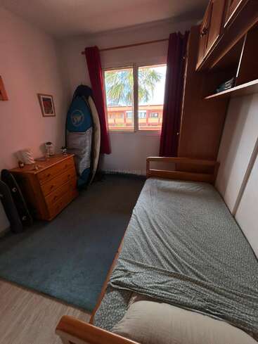 Um pequeno quarto com uma cama de solteiro, lençóis esverdeados, uma cômoda de madeira, um skate, uma prancha de surfe, cortinas vermelhas, uma janela com vista para uma palmeira e um prédio laranja do lado de fora.