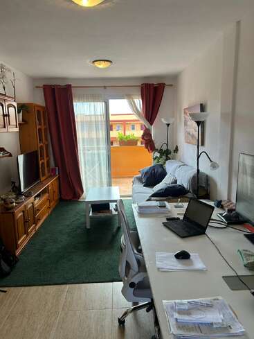 Uma aconchegante sala de estar com uma mesa de trabalho, laptop, papéis, TV, sofá, mesinha de centro, carpete verde, cortinas vermelhas e uma varanda que deixa a luz natural entrar.