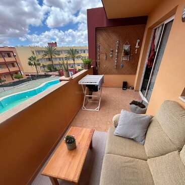 Uma varanda aconchegante com um sofá bege, uma mesa de madeira, um vaso de planta, um varal e decoração na parede tem vista para uma piscina e palmeiras sob um céu azul.