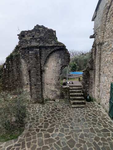 Essa imagem mostra ruínas antigas de pedra com paredes em arco, um pátio pavimentado com pedras, degraus de pedra que levam para cima, árvores nas proximidades, céu nublado e uma piscina azul à vista.