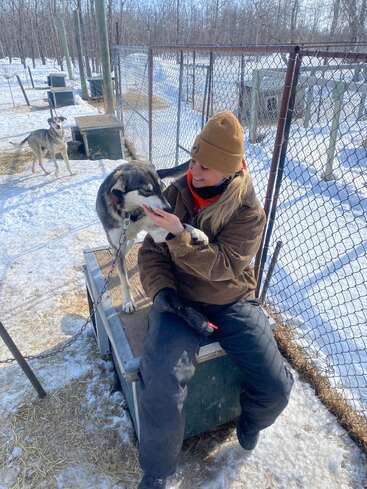Une personne vêtue de vêtements d'hiver est assise sur une niche et sourit en caressant un husky. La neige recouvre le sol et d'autres chiens sont visibles à proximité dans des chenils.