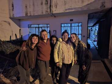 Cinco mulheres estão juntas, sorrindo para a câmera à noite, do lado de fora de um prédio. A iluminação quente destaca suas expressões felizes. O plano de fundo mostra portas e pátios fechados.