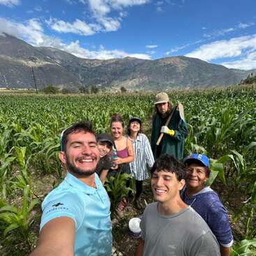 Eine Gruppe von sieben lächelnden Menschen posiert für ein Selfie in einem saftig grünen Maisfeld, umgeben von Bergen und blauem Himmel mit vereinzelten Wolken.
