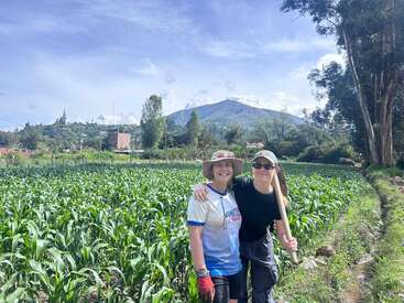 Deux femmes, souriantes, se tiennent côte à côte dans un champ de maïs vert et posent pour une photo. Derrière elles, les montagnes, les arbres et le ciel bleu forment un magnifique décor.