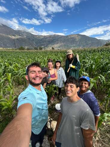 Un groupe de sept personnes souriantes prend un selfie dans un champ de maïs verdoyant, entouré de montagnes et d'un ciel bleu parsemé de nuages.