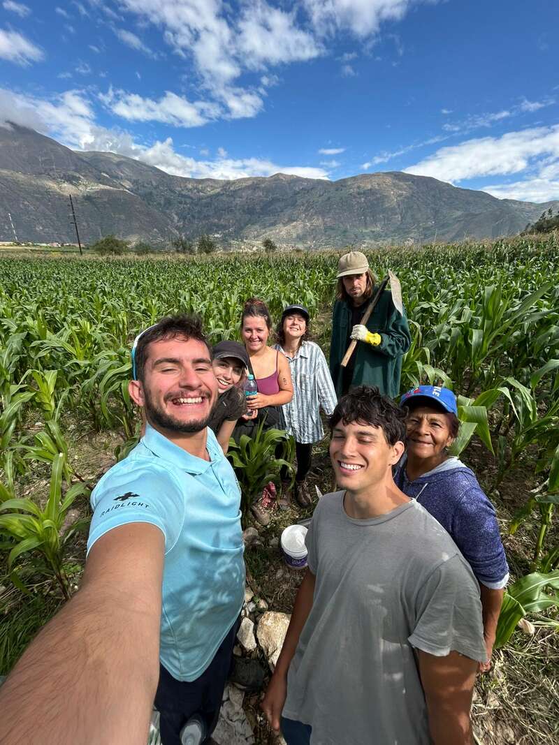 Un groupe de sept personnes souriantes prend un selfie dans un champ de maïs verdoyant, entouré de montagnes et d'un ciel bleu parsemé de nuages.