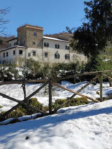 Ein großes, rustikales Gebäude steht inmitten einer verschneiten Landschaft, umgeben von Bäumen und einem Holzzaun, über ihm ein strahlend blauer Himmel und winterliche Sonne.