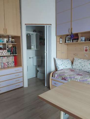 This cozy bedroom features a single bed, built-in wooden cabinets, bookshelves, a small table, and an open door revealing a compact bathroom with a toilet.