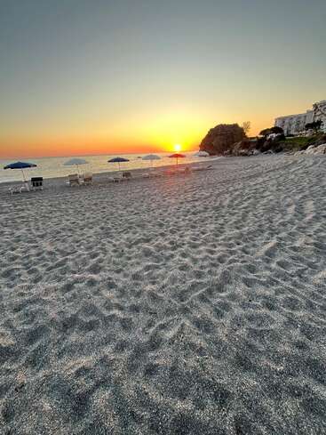 Golden sunset over a tranquil beach with empty sun loungers and umbrellas, gentle waves, textured sand, rocky cliffs, and white buildings under a clear sky. Peaceful scene.