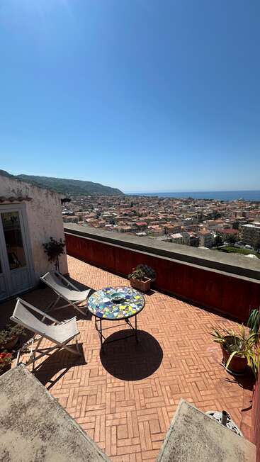 A sunny rooftop terrace with two lounge chairs and a colorful mosaic table overlooks a charming town, hills, and the distant blue sea under a clear sky.
