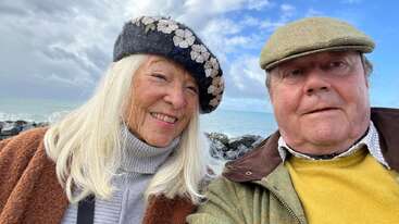 An elderly couple stands outdoors by the sea, smiling warmly. Both wear hats and cozy clothing. The sky is partly cloudy, creating a peaceful, joyful scene.