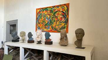 Seven sculpted busts are displayed on a white table beneath a vibrant, abstract painting with swirling patterns. Firewood is stacked below, creating an eclectic atmosphere.
