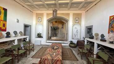 This elegant room features classical busts, vibrant artwork, a patterned rug, green chairs, a central table, and a grand arched entryway with stairs and sculpture.