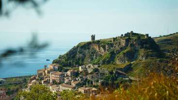 A picturesque coastal village nestles below a dramatic hilltop castle overlooking the serene blue sea, surrounded by greenery and sunlit Mediterranean architecture under a clear sky.