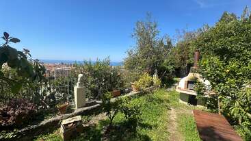 A sunlit garden with lush greenery, potted plants, and a stone barbecue overlooks a scenic coastal town and the blue sea under a clear sky.