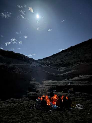 A group of people sit around a glowing campfire at night, surrounded by rocky hills under a bright full moon and scattered clouds in a serene atmosphere.