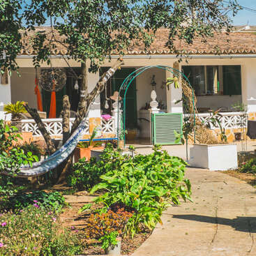 L'image représente une maison blanche sereine avec un toit en tuiles, entourée d'une verdure luxuriante et de plantes éclatantes, dégageant un sentiment de tranquillité et de beauté naturelle.