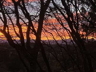 Bare tree branches silhouette a vibrant, colorful sunset of reds, oranges, and purples. Hills and scattered distant lights are visible beneath the dramatic sky.