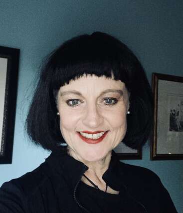 A woman with short black hair and bangs smiles brightly at the camera. She wears red lipstick, pearl earrings, and a black top. Framed art hangs behind.