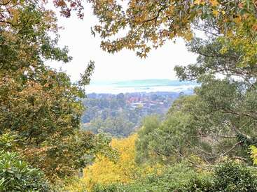 A scenic view framed by leafy trees with autumn colors. In the distance, a town nestles among hills, with a lake and hazy mountains beyond.