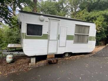 A white and green vintage caravan is parked on a paved area beside trees. It has two windows with curtains, a gas bottle, and sits on bricks.