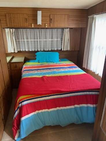This image shows a cozy bedroom with wood-paneled walls, a colorful striped bedspread, blue pillow, curtained windows, and small cabinets above and beside the bed.