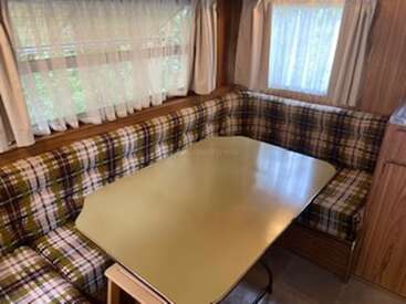 This image shows a cozy RV dining area with plaid cushioned benches, a yellow rectangular table, wood-paneled walls, and curtains covering large windows, creating a retro vibe.