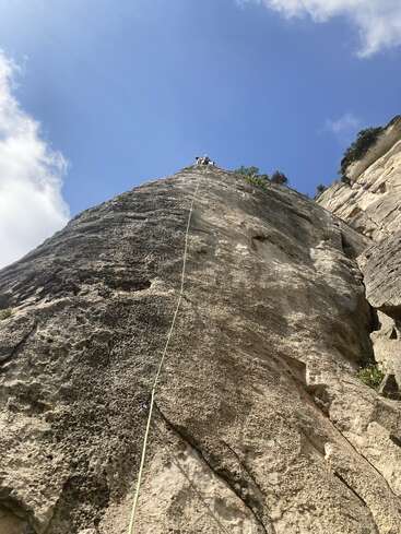 A rock climber reaches the top of a steep, textured cliff, secured by a rope, under a bright blue sky with scattered white clouds.