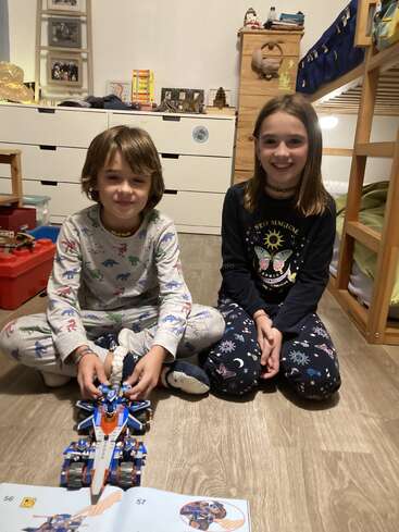 Two children in pajamas sit on a bedroom floor, smiling. They are playing with a blue toy vehicle, with assembly instructions and bunk beds visible behind them.