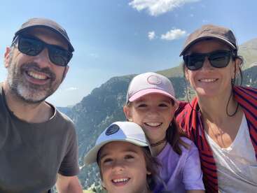A smiling family of four, wearing hats and sunglasses, enjoys a sunny day outdoors. Mountains and a bright blue sky with clouds form the scenic backdrop.