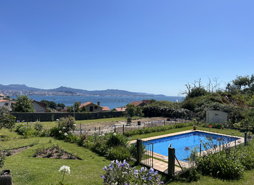 A beautiful backyard with a fenced swimming pool, lush green grass, flowers, and a gorgeous view of houses, mountains, and a sparkling blue sea.