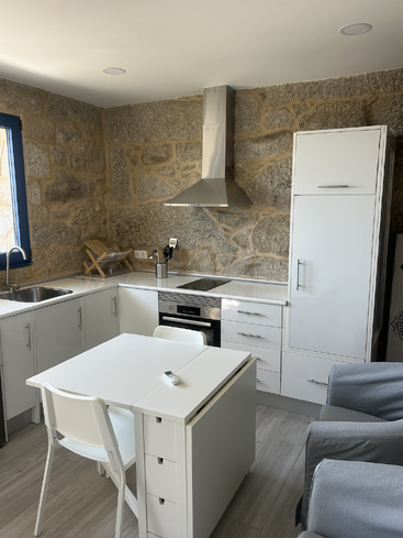 This image shows a modern kitchen with stone walls, white cabinets, a small dining table, stainless steel appliances, a sink, and comfortable gray chairs. Cozy atmosphere.