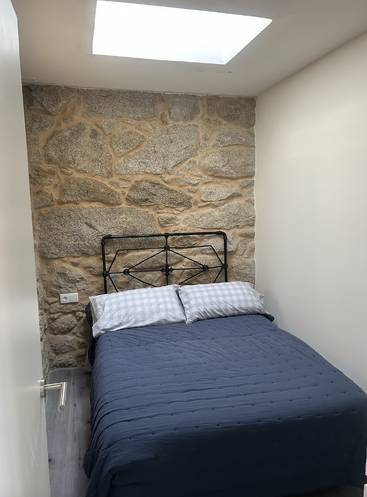 A cozy bedroom features a stone accent wall, metal headboard, blue bedspread, checkered pillows, skylight above, white walls, and a wooden floor. Simple design.