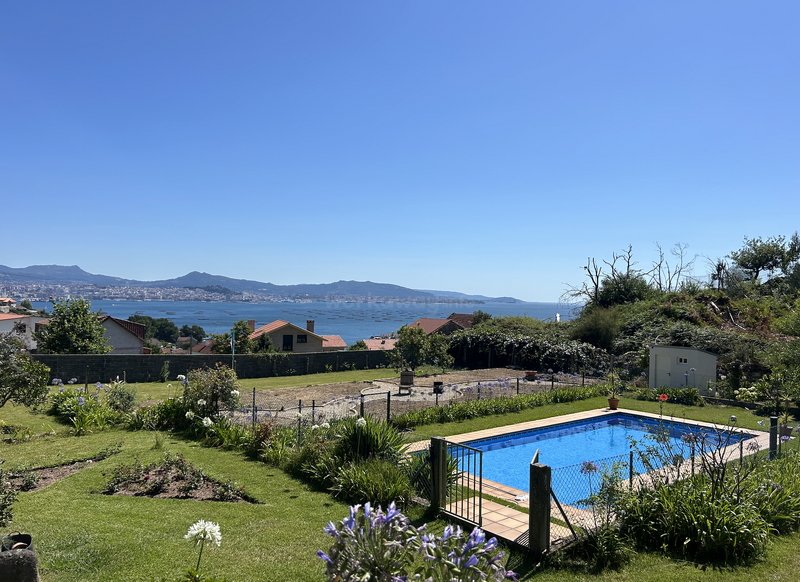 A beautiful backyard with a fenced swimming pool, lush green grass, flowers, and a gorgeous view of houses, mountains, and a sparkling blue sea.