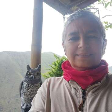 A imagem mostra uma mulher de cabelos curtos, vestindo uma jaqueta bege e um lenço vermelho, ao lado de uma estátua de coruja, em um cenário montanhoso.