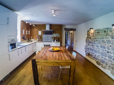 La imagen muestra una cocina moderna con armarios y encimeras blancas, una mesa de comedor de madera con sillas y una pared de piedra al fondo.