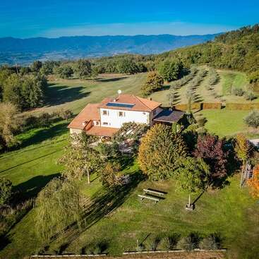 La imagen representa un sereno paisaje rural con una casa de dos plantas con tejado de terracota, rodeada de frondosos árboles y una vasta extensión de verdes campos bajo un cielo azul despejado.