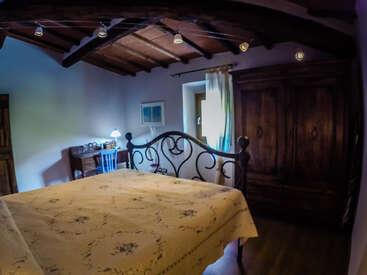 La imagen muestra un dormitorio sereno con un somier de madera oscura, un armario de madera y un escritorio, con una pared blanca y un techo de madera con vigas vistas.