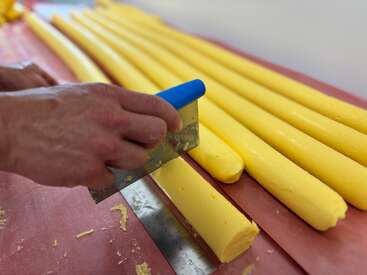 A person is cutting long, yellow cylindrical pieces of dough or candy with a metal scraper, creating evenly sized segments on a red surface in a kitchen.
