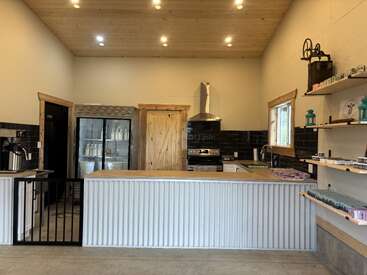 This cozy shop features a wooden ceiling, modern lighting, a corrugated counter, open shelving with soaps, a refrigerator, and stainless steel appliances for a rustic, welcoming vibe.