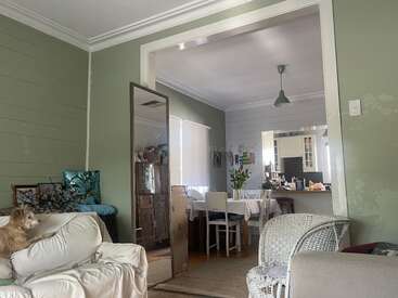 Uma sala de estar aconchegante com paredes verdes, acabamento branco, móveis de vime e estofados, um cachorro pequeno em uma cadeira e vista para uma cozinha iluminada pelo sol.