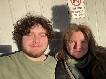 Um jovem e uma mulher estão sentados juntos, sorrindo à luz do sol. Atrás deles há uma parede com uma placa que diz: "Proibido comer ou beber no campo".